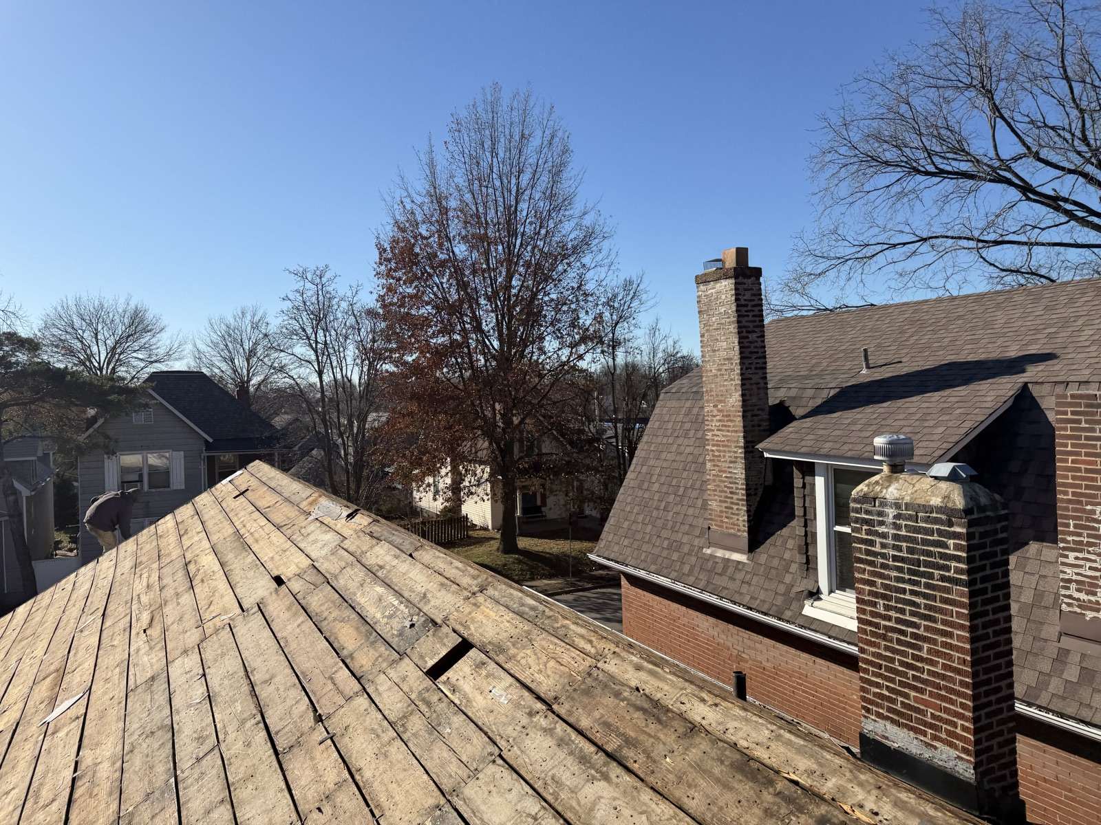 A hip roof stripped to bare plank decking in the foreground with gaps open between the boards, a brick chimney and a brown shingled roof still intact behind it, and bare winter trees over the neighbouring houses