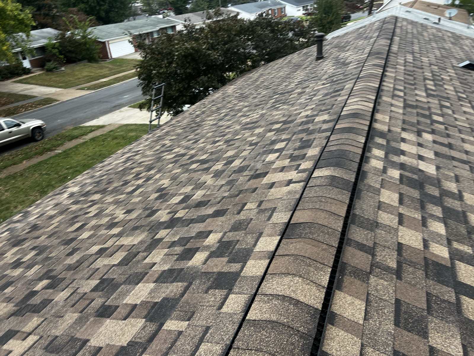 Looking straight down the ridge of a finished brown and tan multi tone shingle roof, with the ridge cap shingles curving over a black continuous ridge vent, a ladder leaning against the eave and a street of single storey suburban houses below