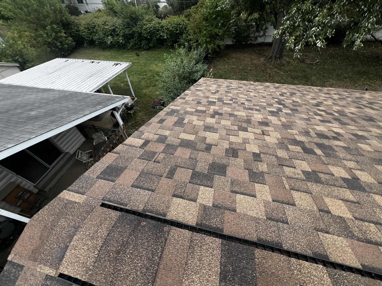 An overhead view of a finished brown and tan architectural shingle roof plane meeting the ridge, with the black ridge vent visible under the cap, and a back garden, a white metal patio cover and a charcoal grill below