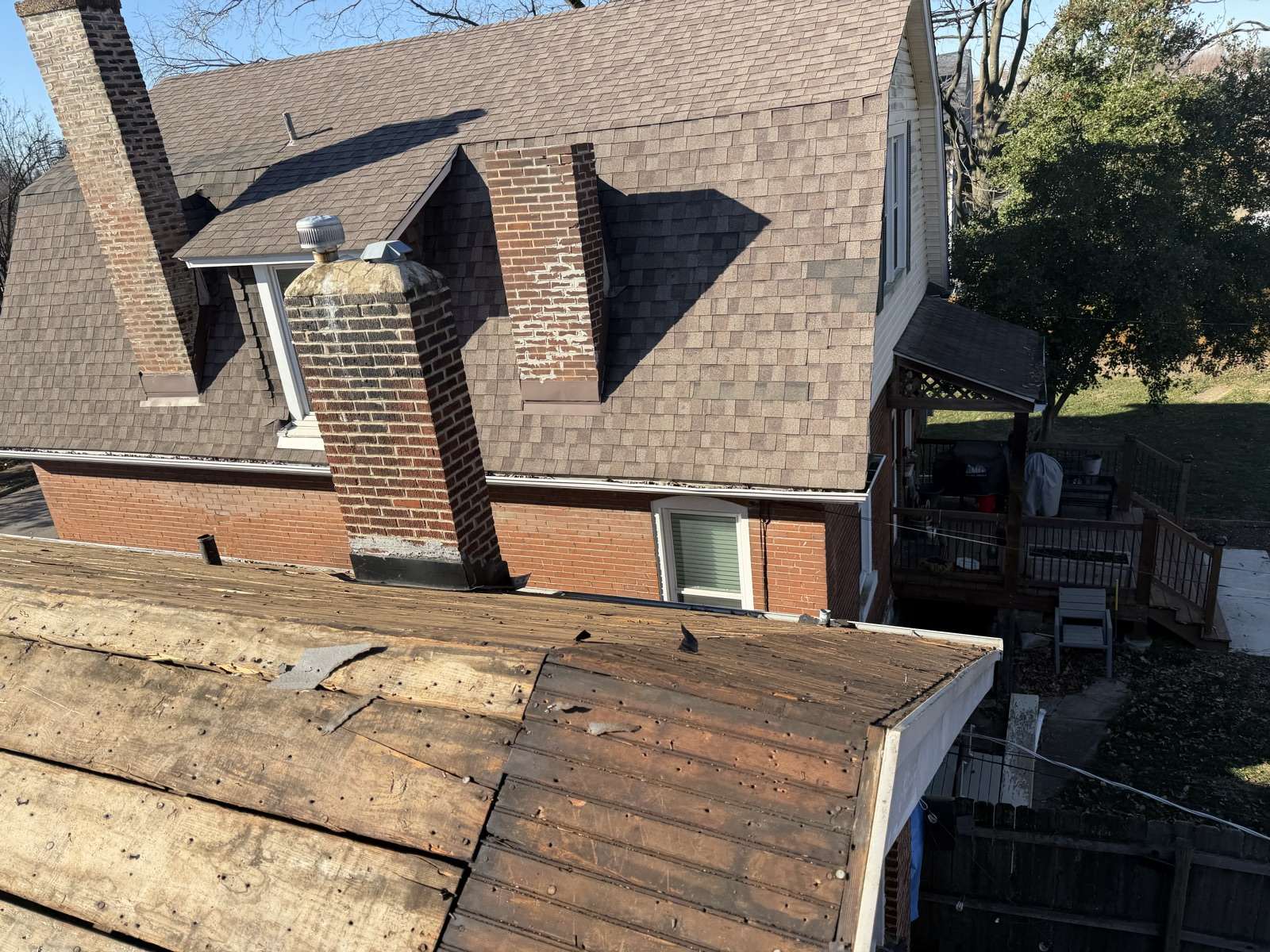 A brick house with brown shingles still on the upper roof, while the lower porch roof in the foreground has been stripped back to bare board decking with old nail heads and torn felt still showing, and the chimney brickwork visibly spalled
