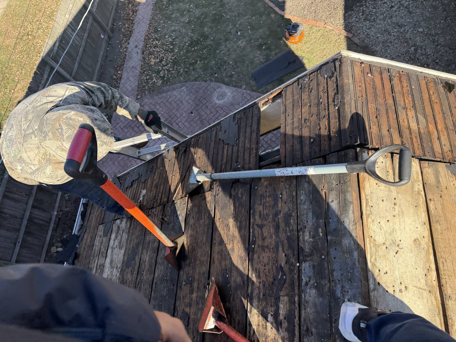 Looking down from above at a porch roof stripped to dark weathered plank decking, with two tear off spades and a pry bar lying on the boards and a roofer in a camouflage jacket kneeling at the edge beside a ladder