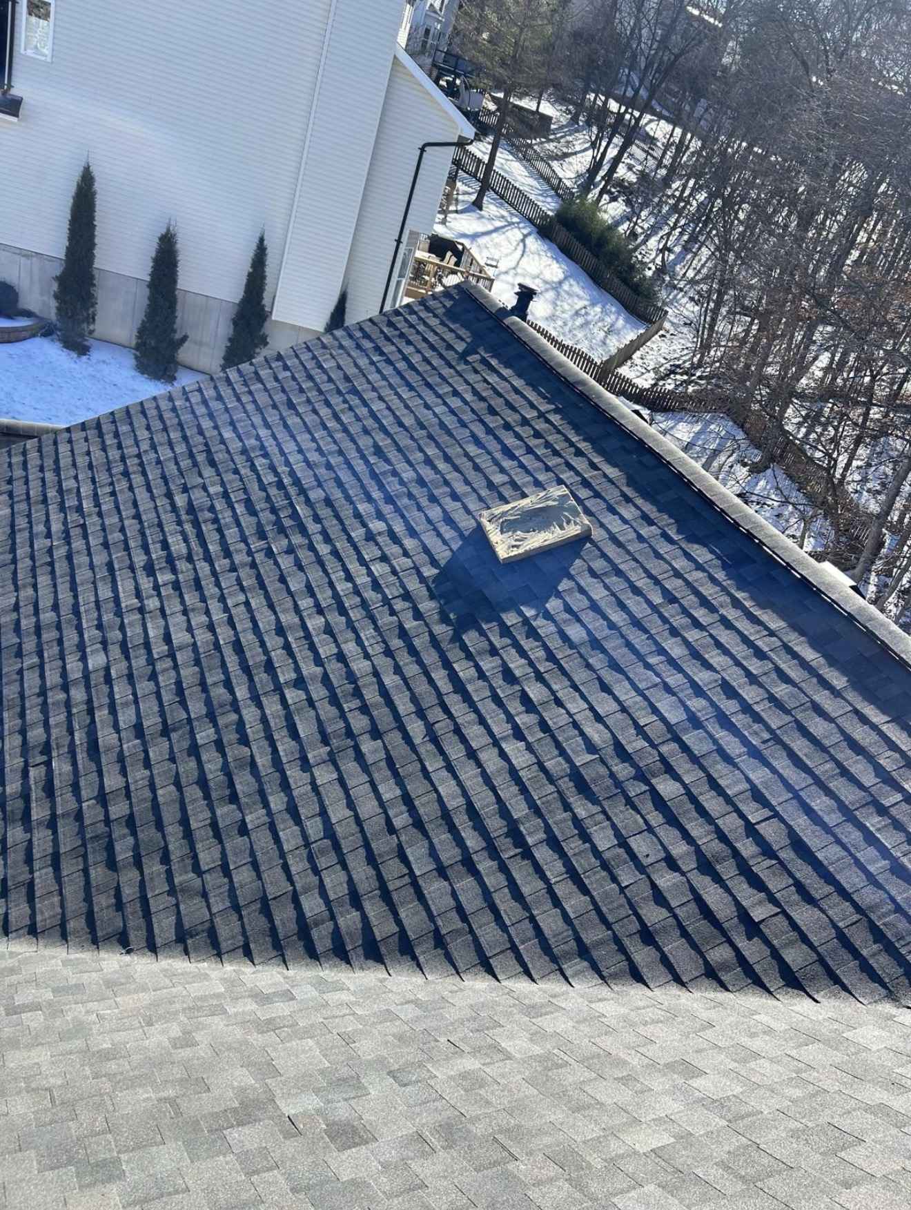 A steep charcoal grey shingle slope lit by low winter sun so every course throws a hard shadow line, with a scrap offcut left lying on the slope, snow on the ground and a neighbouring white sided house a few feet away